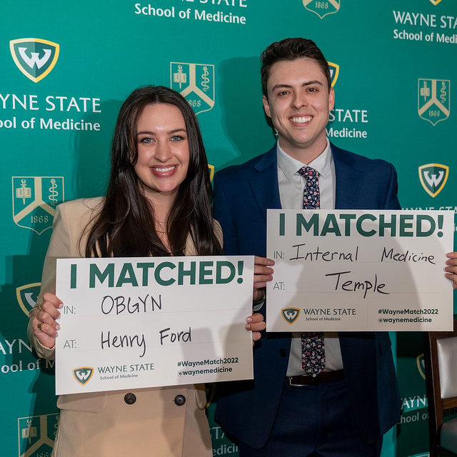 Male and female medical students smiling and standing next to each other, both holding signs that read