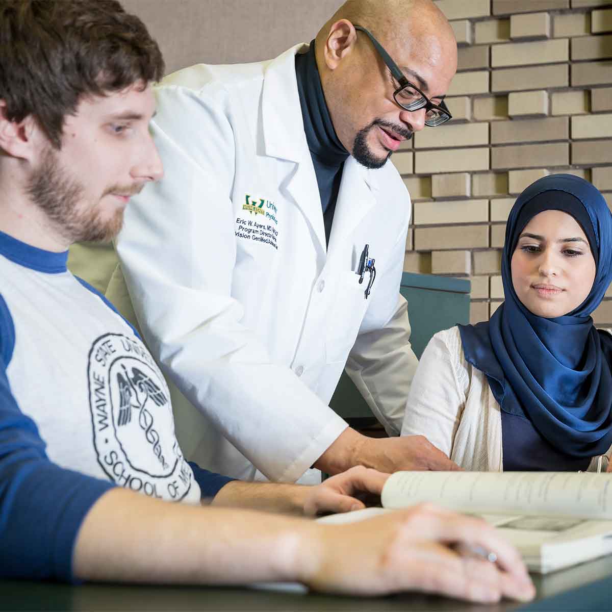 Male professor wearing white coat bending over to talk with male and female students, who are both sitting down at a desk looking at an open book.
